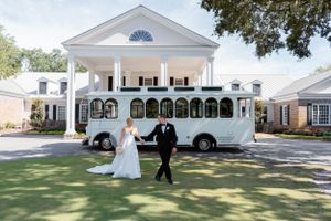 Elegant wedding party poses outside white columned mansion with vintage trolley bus in the background.