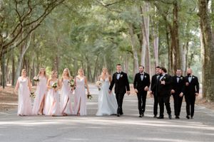 Wedding party walks along a tree-lined path with bridesmaids in pink and groomsmen in black.