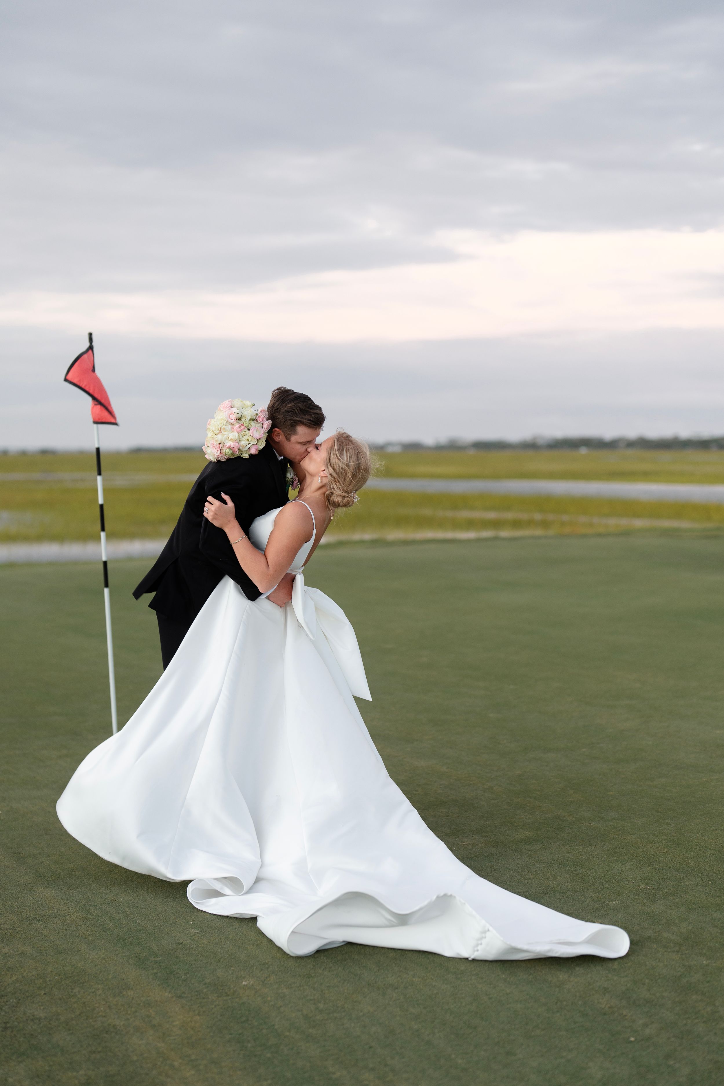A romantic wedding photo shows a couple embracing on a golf course with a flowing white dress and red flag pin.