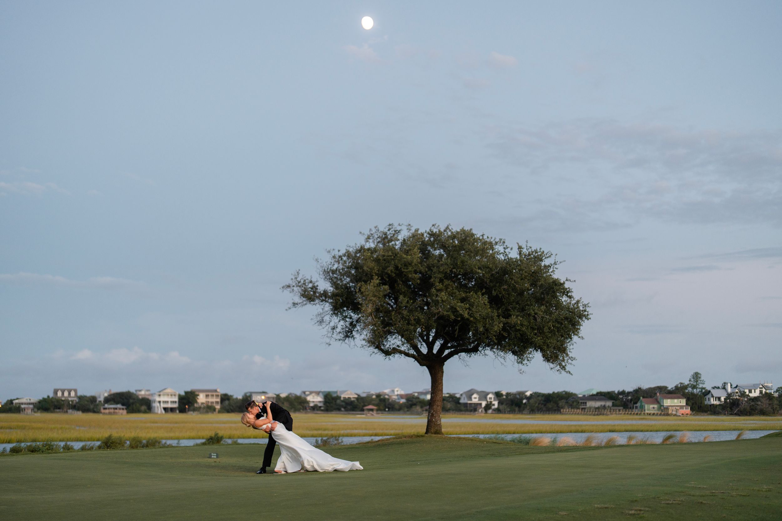 A lone tree stands on a golf course with a couple in formal attire posing against a dusky sky with visible moon.