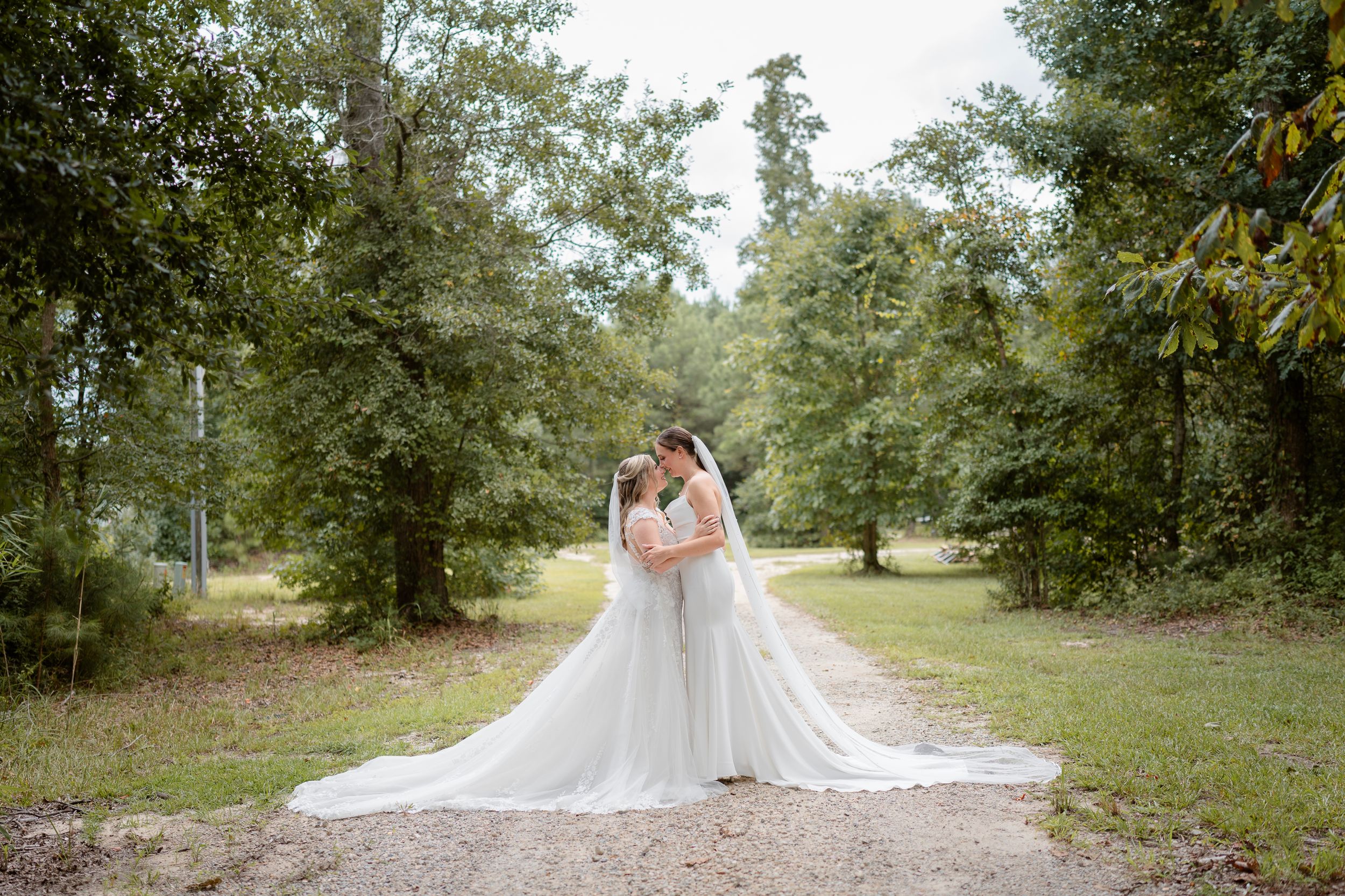 Two people share an intimate moment on a wooded path wearing flowing white wedding dresses in an outdoor setting.