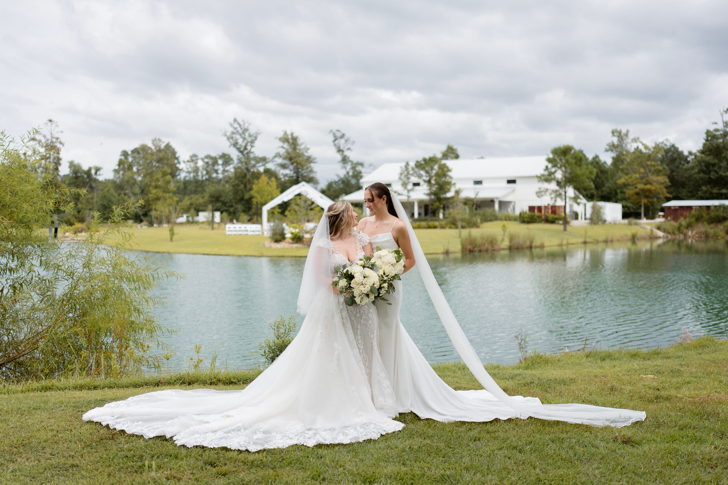 Elegant wedding scene by a serene lake with flowing white gown and bouquet against cloudy sky.