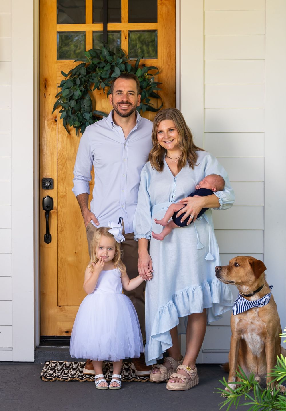 Family portrait on porch with brown dog and wooden door showing casual home lifestyle.