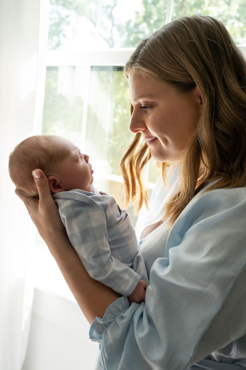 Tender moment with newborn in blue clothing near sunlit window.
