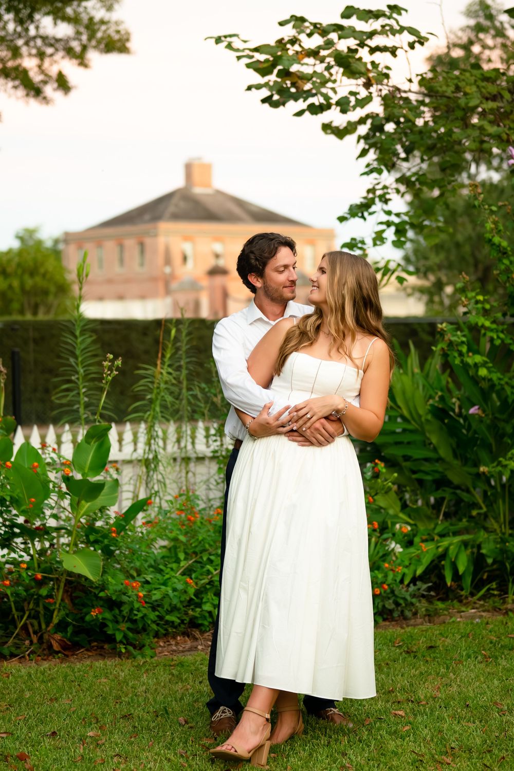 A romantic couple in white outfits embrace and laugh together in a lush garden setting with buildings in the background.