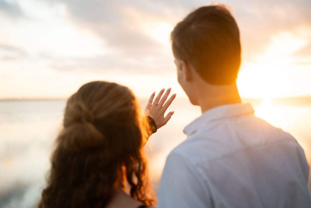 Silhouetted couple watches golden sunset over calm water as sun creates warm backlit glow.