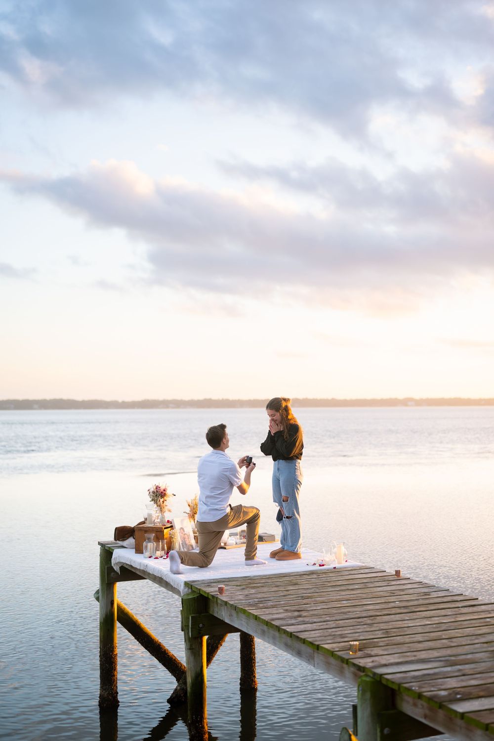 A couple sits on a wooden pier overlooking a serene lake at sunset with a picnic spread beside them.