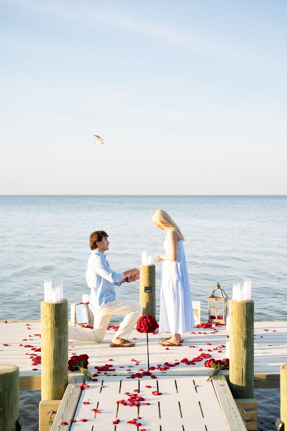 Man proposes on rose petal-covered dock by the ocean, woman in white dress, candles and red roses surrounding them.