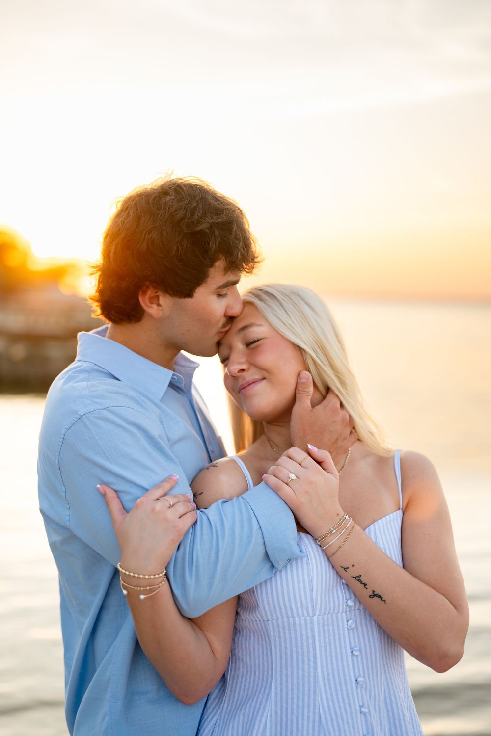 Couple embracing at sunset on a beach, man kissing woman's temple, both wearing light blue outfits.