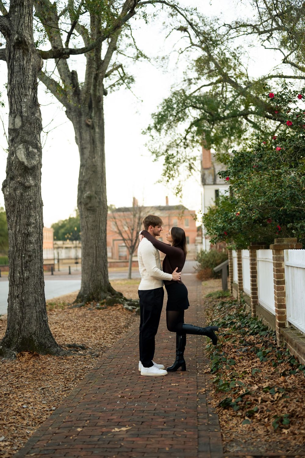A romantic couple dances together on a brick pathway surrounded by autumn leaves and tall trees.