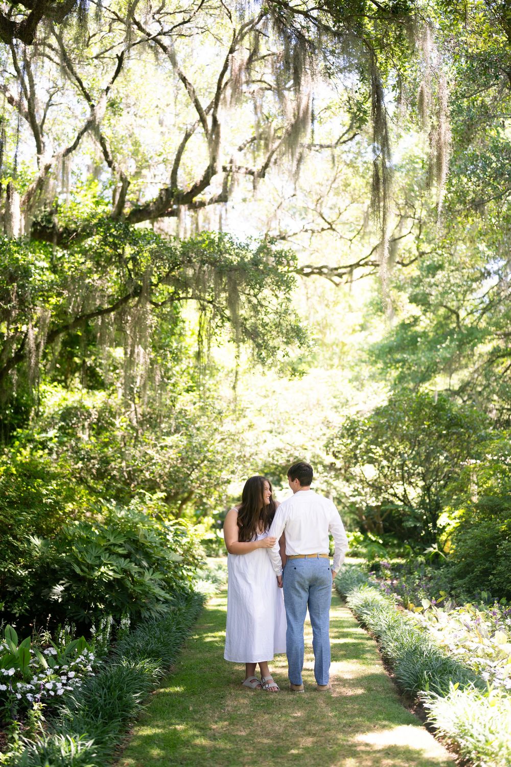 Couple walking hand in hand down a lush garden path beneath sprawling moss-draped oak trees.