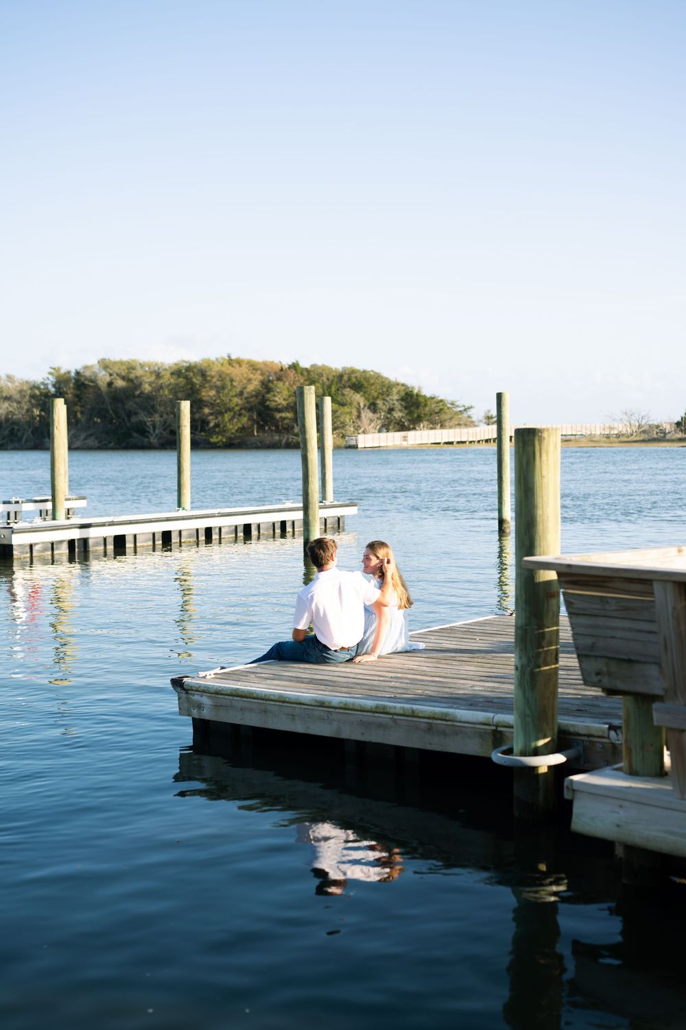Couple sitting together on a wooden dock over calm water, enjoying a peaceful waterfront view on a clear sunny day.