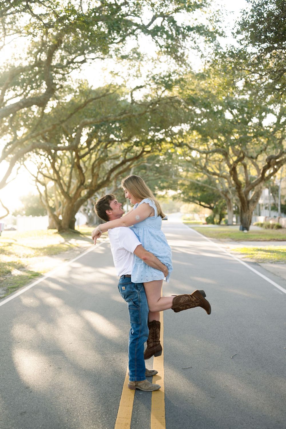 Man lifting woman in his arms on a tree-lined road during golden hour engagement photo session.