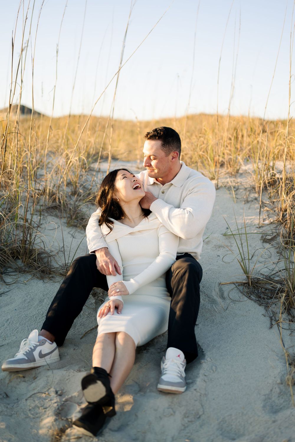 Romantic couple in white outfits share tender moments while sitting on sandy beach dunes at sunset.