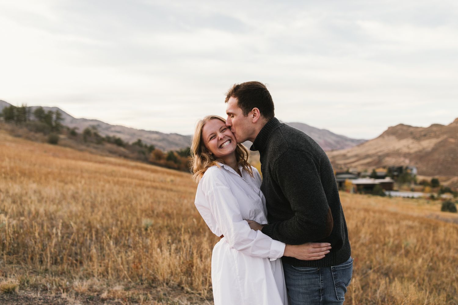 Couple embracing and laughing together in golden field with mountains in background.