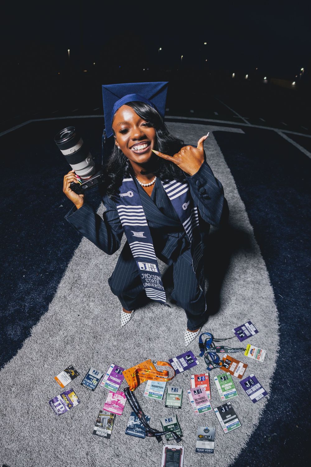Graduate in cap and gown holding camera, posing with collection of event passes and lanyards spread on ground at night.