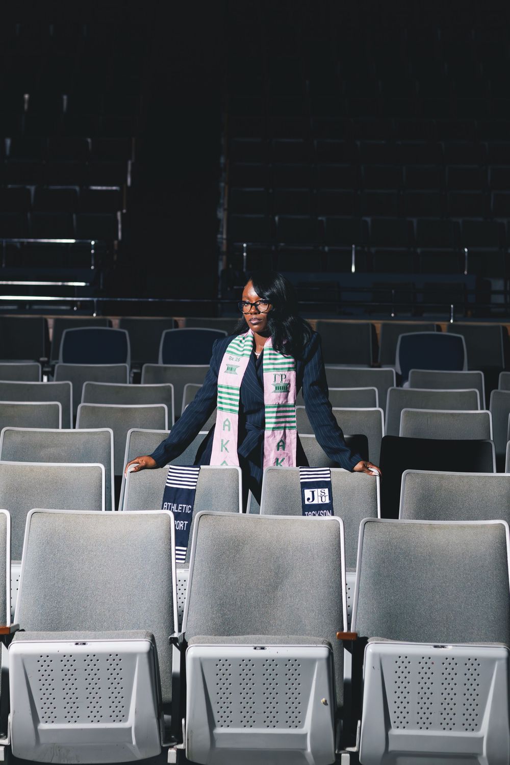 Woman wearing a colorful scarf stands among rows of empty gray auditorium seats in a dark venue.