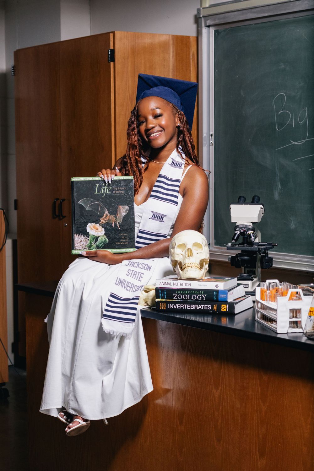 Graduate poses in cap and gown with biology textbooks and skull in a Jackson State University classroom.