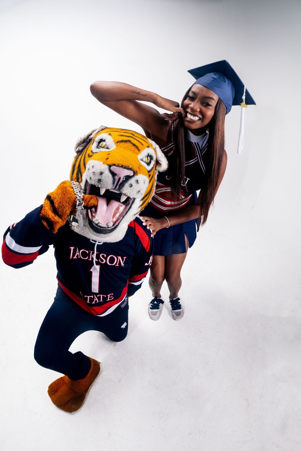 Jackson State University graduate poses with tiger mascot in cap and gown, cheerleader uniform, smiling joyfully.