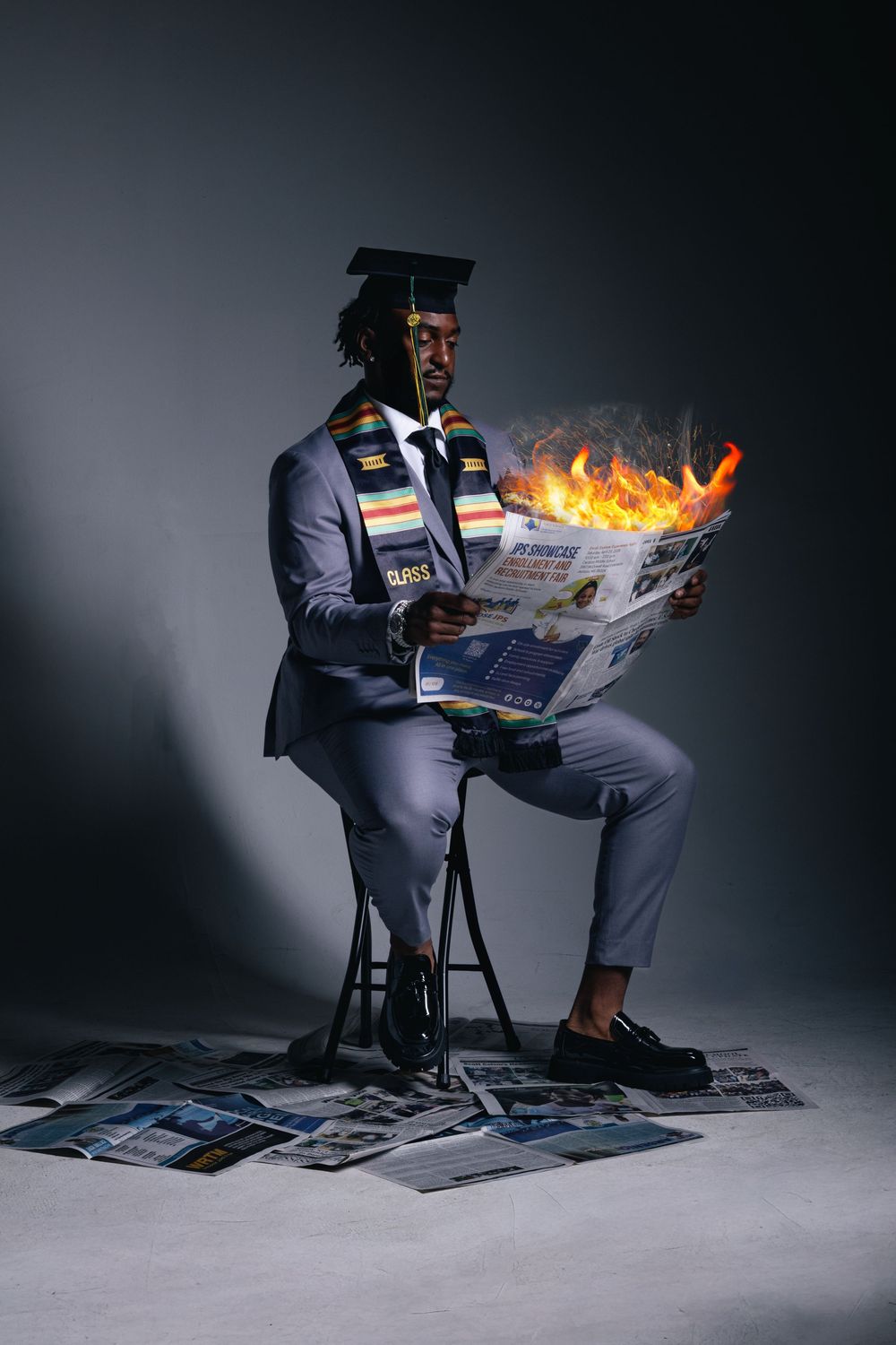 Graduate in cap and stole reads burning newspaper while seated, surrounded by papers on studio floor.