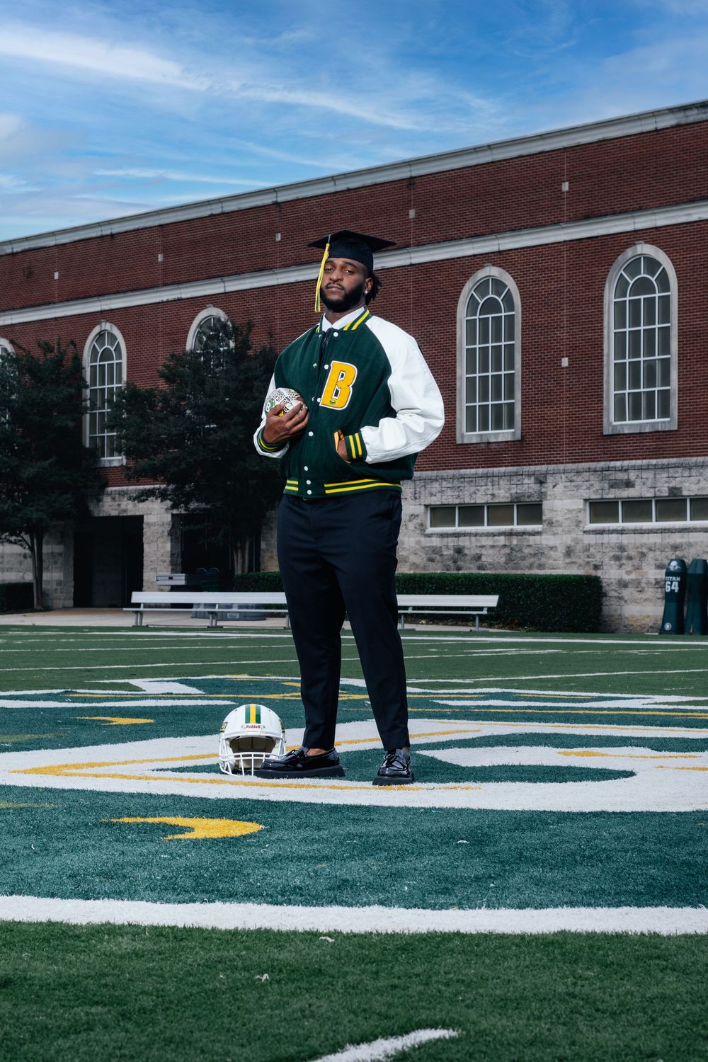 A graduating student in a cap and varsity jacket stands on a football field holding a helmet outside a brick building.