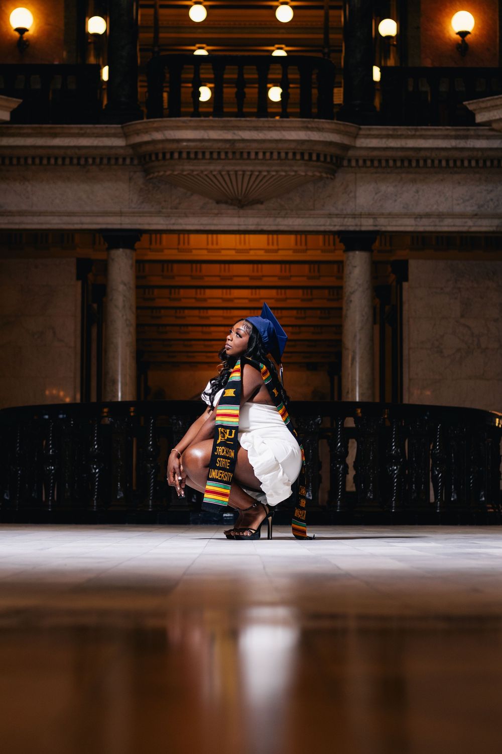 Woman in white outfit and colorful sash crouches gracefully in an elegant columned hall with warm ambient lighting.