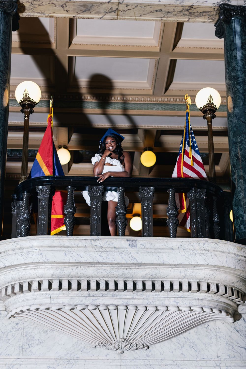 Graduate in white dress and blue cap stands on ornate marble balcony with Arizona and US flags.