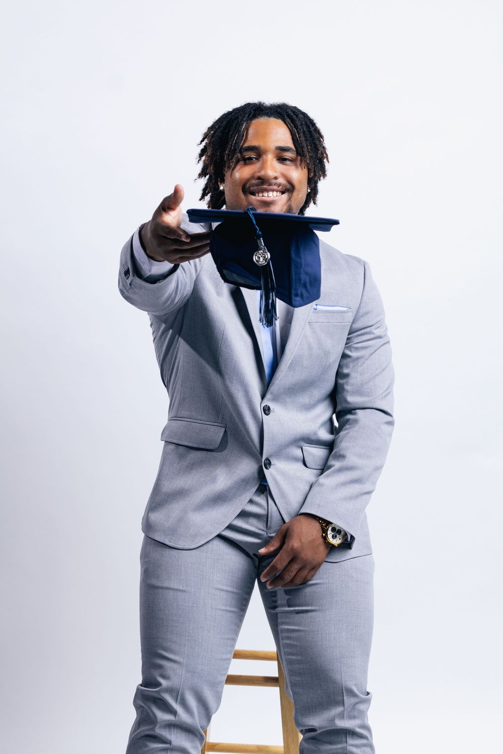 Smiling young man in gray suit holding blue graduation cap toward camera, seated on stool against white background.