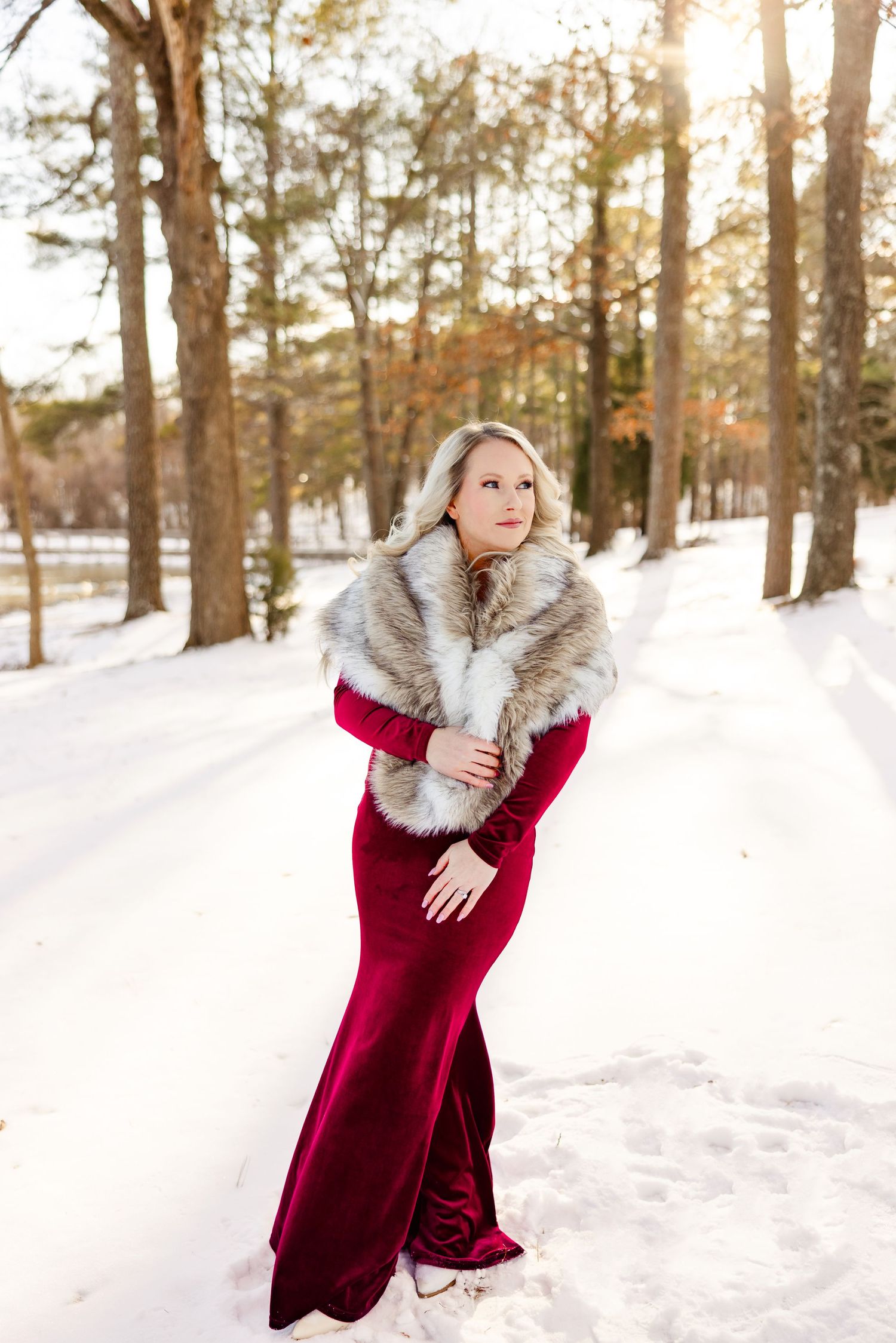 A woman in a red velvet gown and fur stole poses in a snowy forest during golden hour.