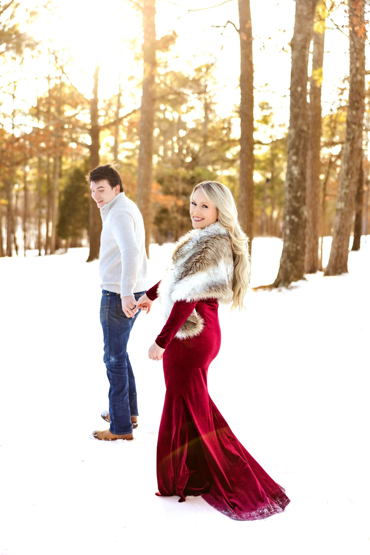 Couple poses in snowy forest setting with woman wearing elegant red velvet dress and fur stole.