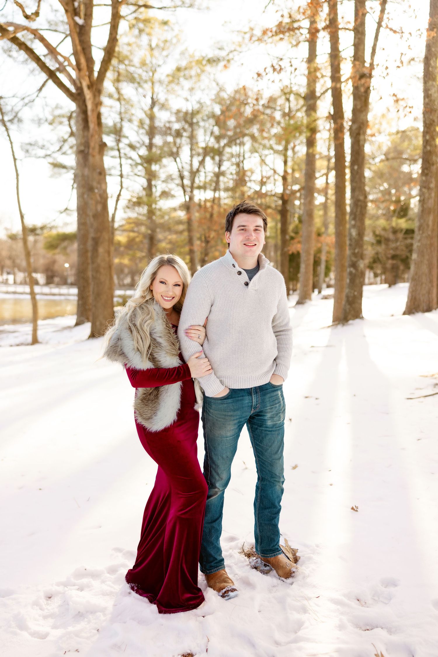 A couple stands together in a snowy winter setting, with one wearing a burgundy dress and the other in a light sweater.