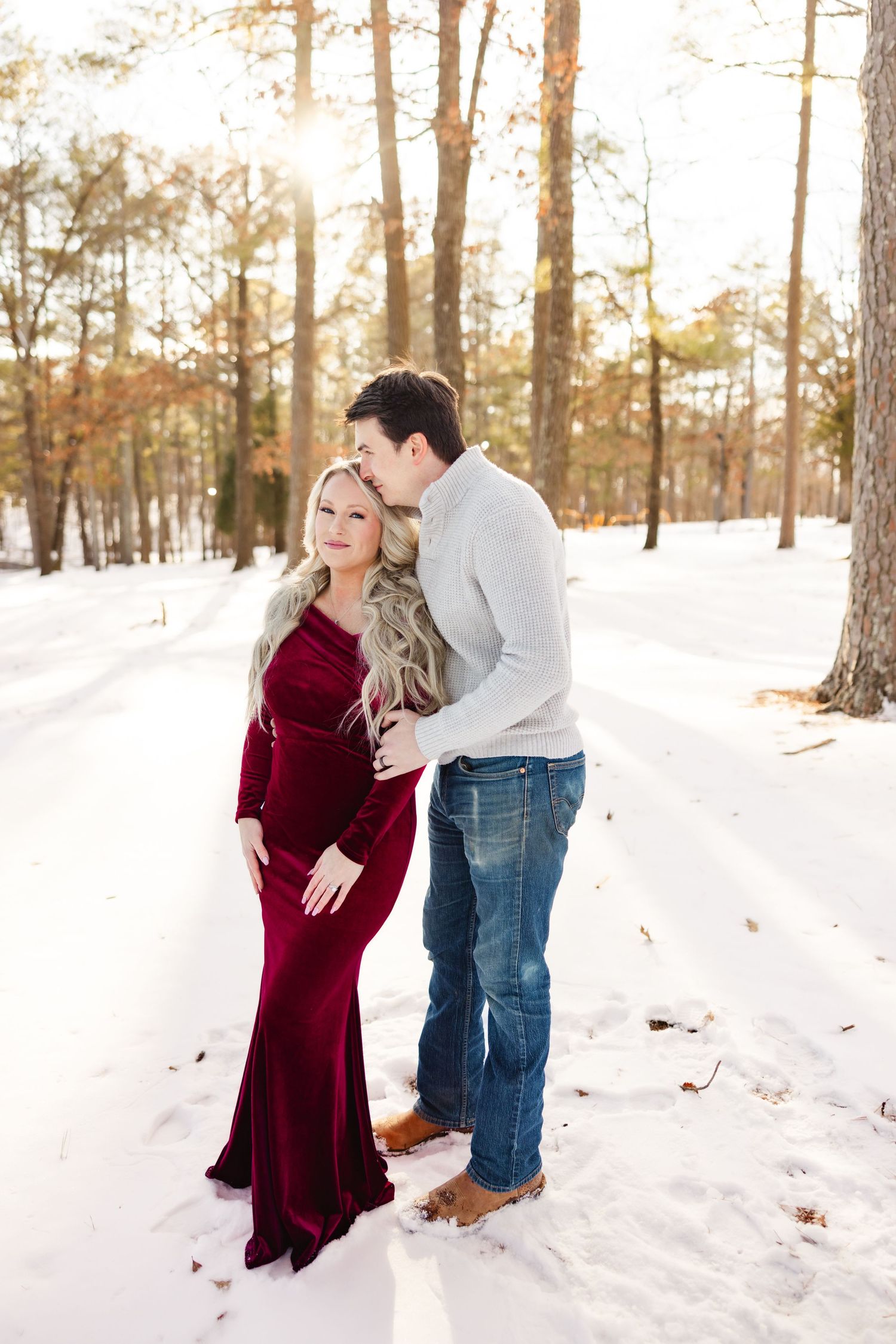 A couple embraces in a snowy winter forest scene, with the woman wearing a stunning burgundy velvet gown.