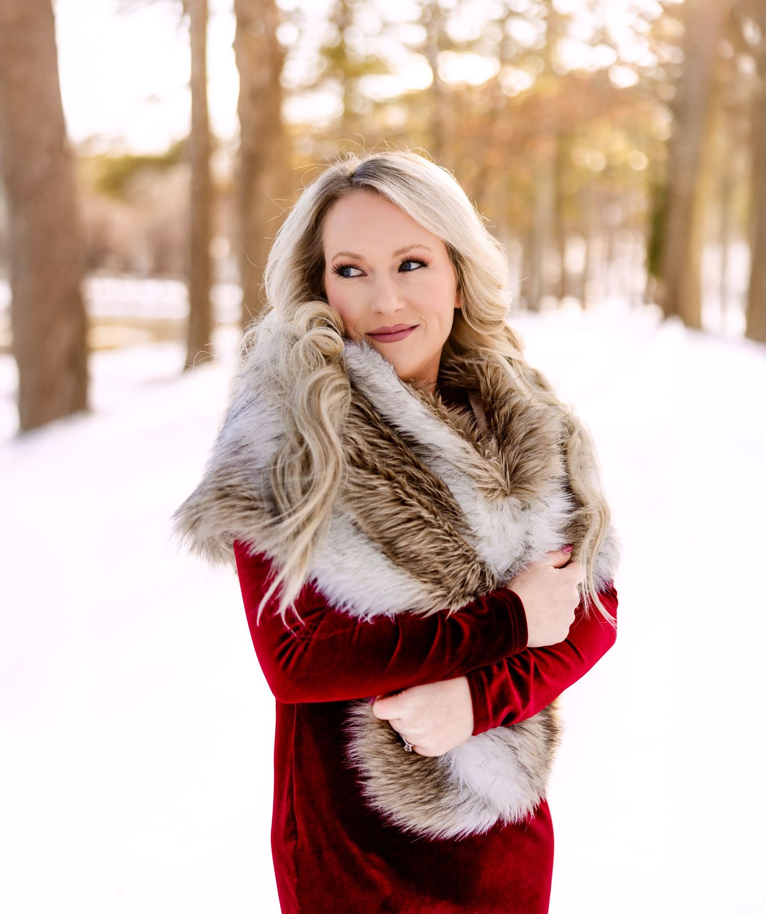 A blonde woman in a red velvet dress and fur stole poses in a snowy forest setting at sunset.