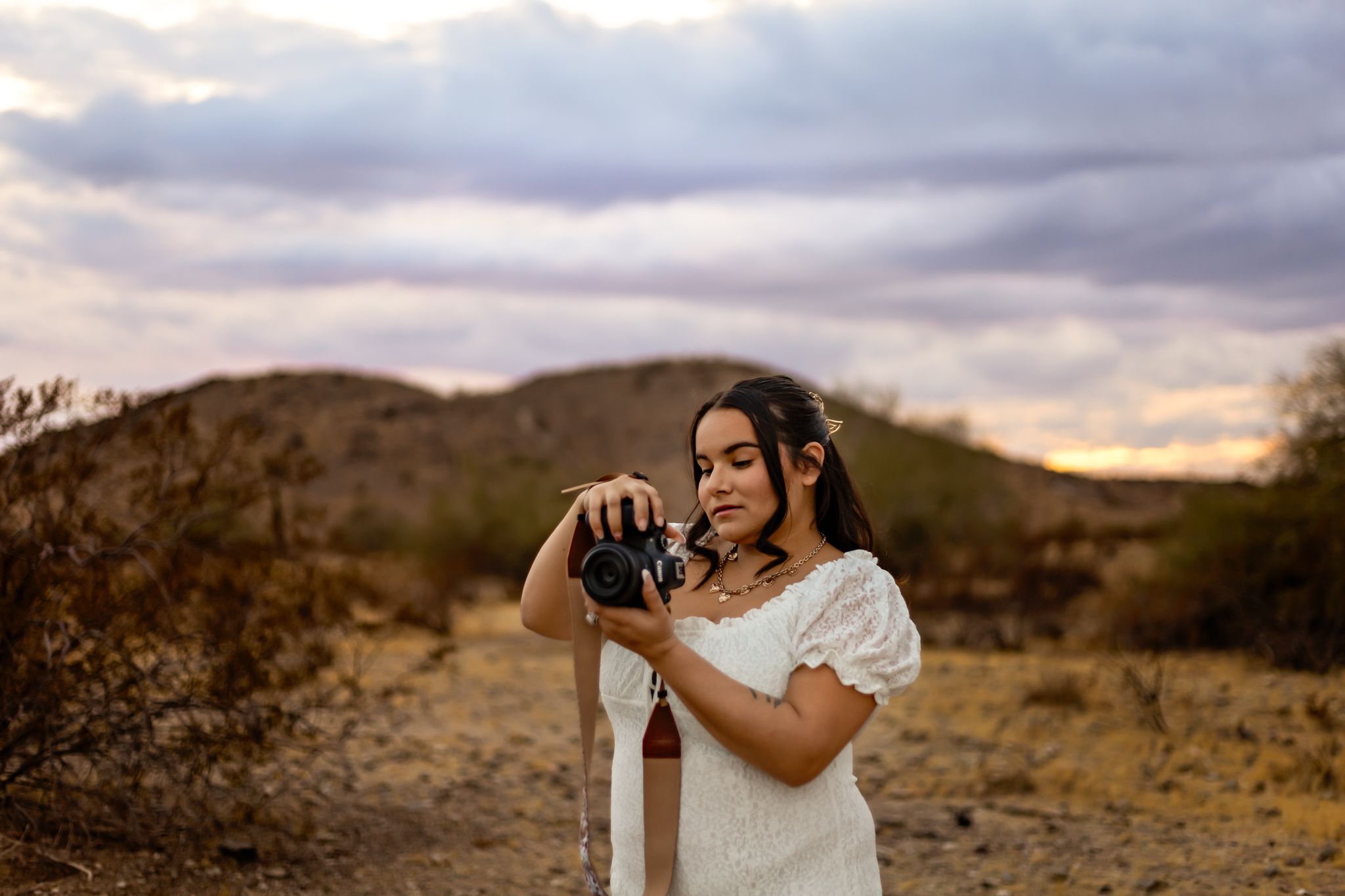 Woman in white dress taking photos in desert landscape at dusk.