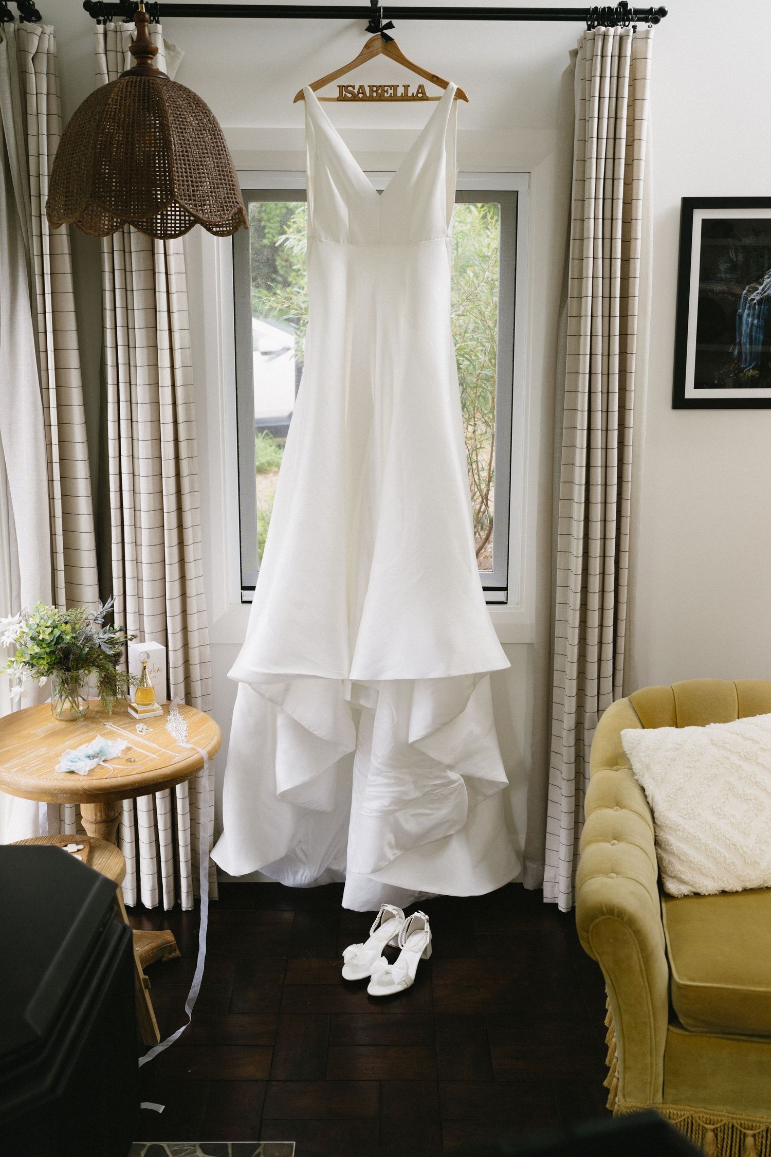 An elegant white wedding dress hangs by a window with white shoes beneath.