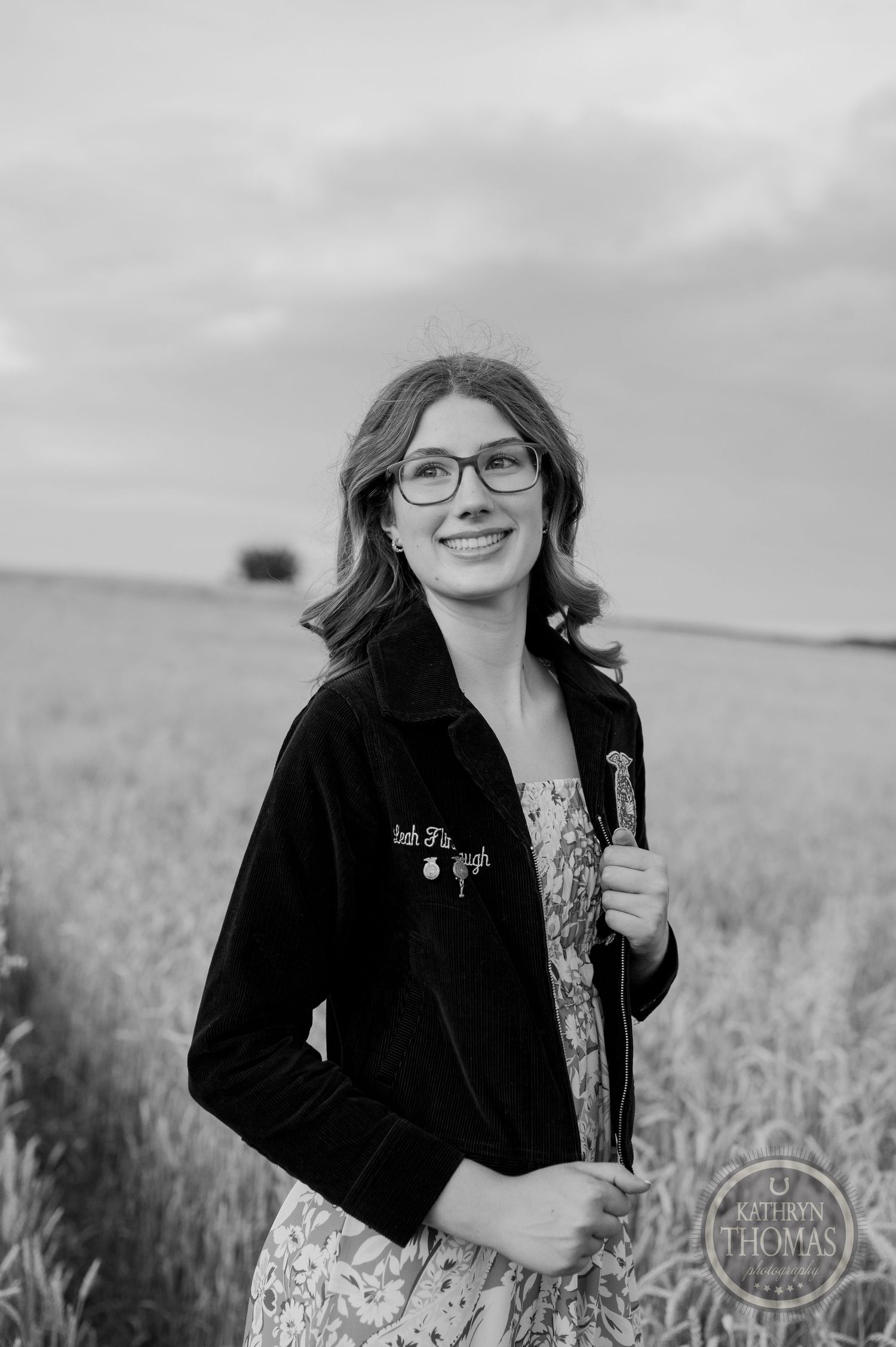 Black and white portrait of someone in an FFA jacket and floral dress standing in a wheat field.