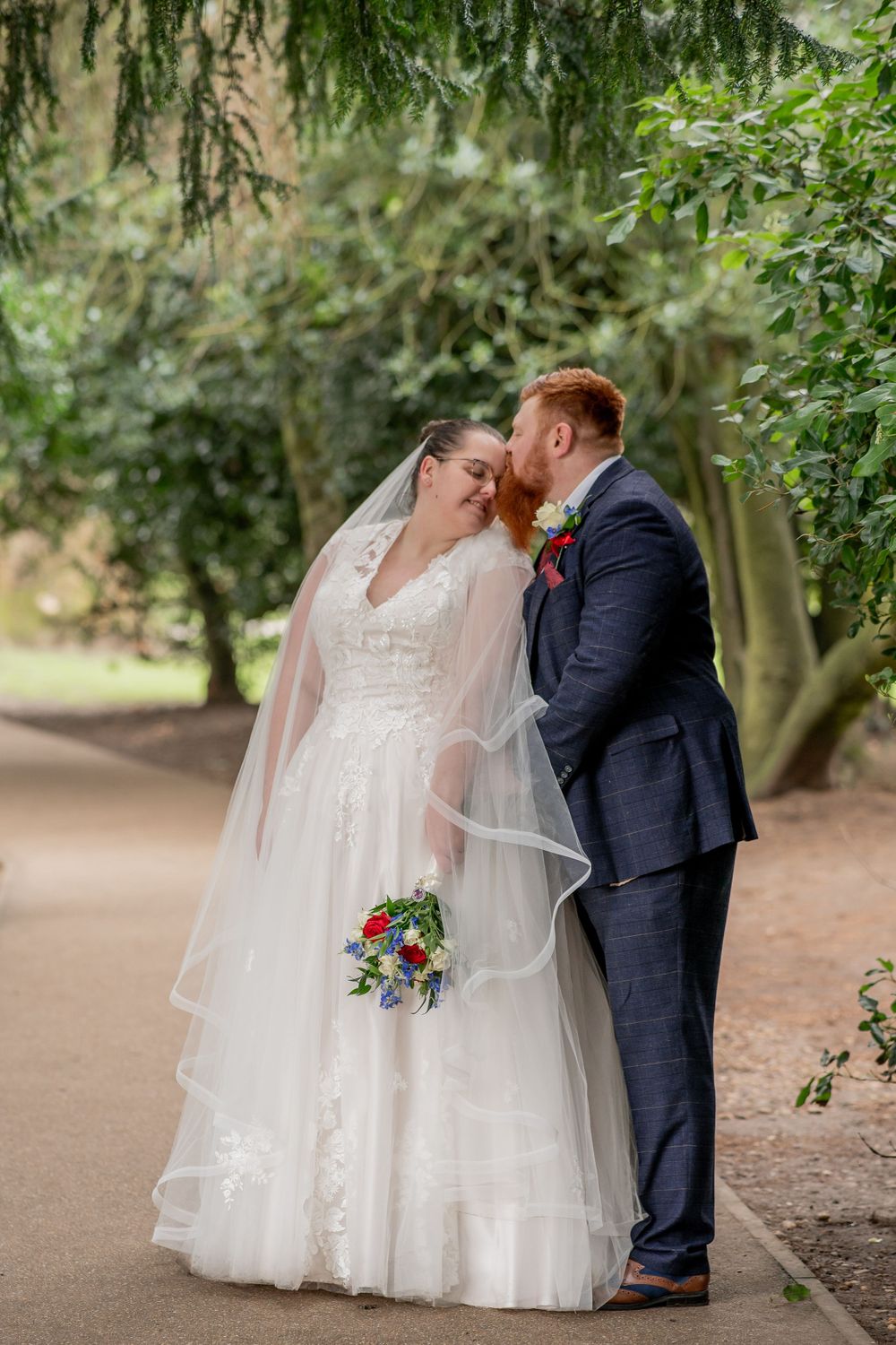 Newlyweds share an intimate moment on a tree-lined garden path with colorful bridal bouquet.