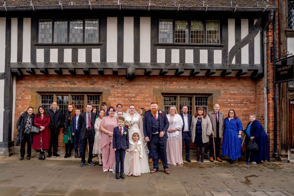 Wedding party poses together outside historic Tudor-style building with timber frame and brick walls.
