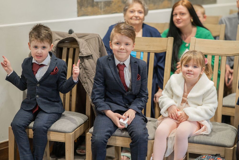 Three children in formal attire seated on wooden chairs at a wedding with adults in background.