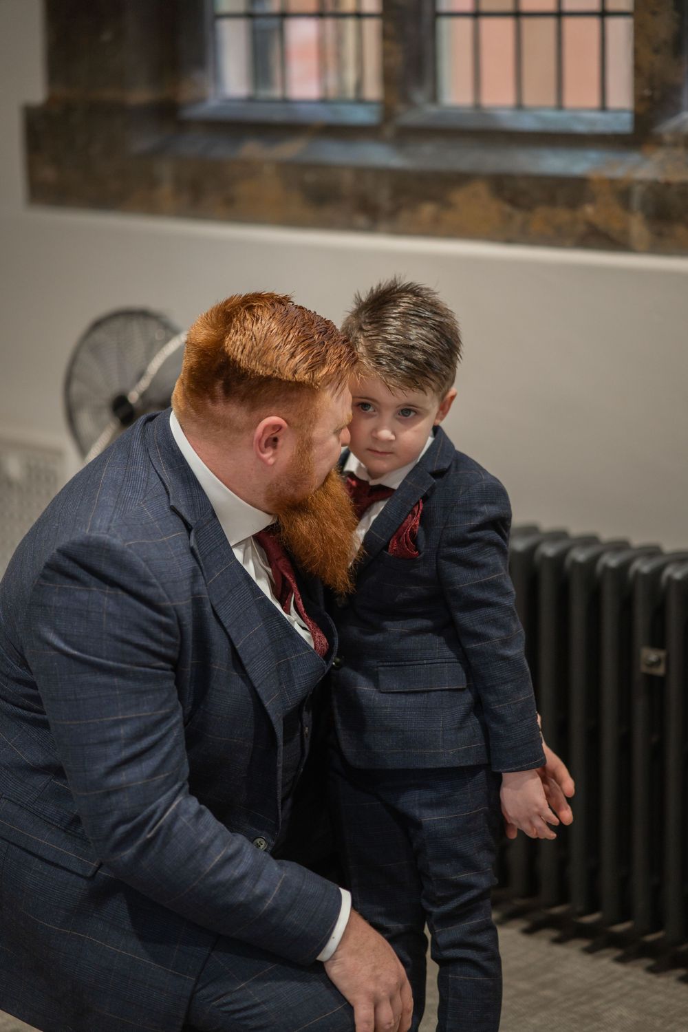 A tender moment shared between two people in matching navy suits standing next to a vintage radiator indoors.