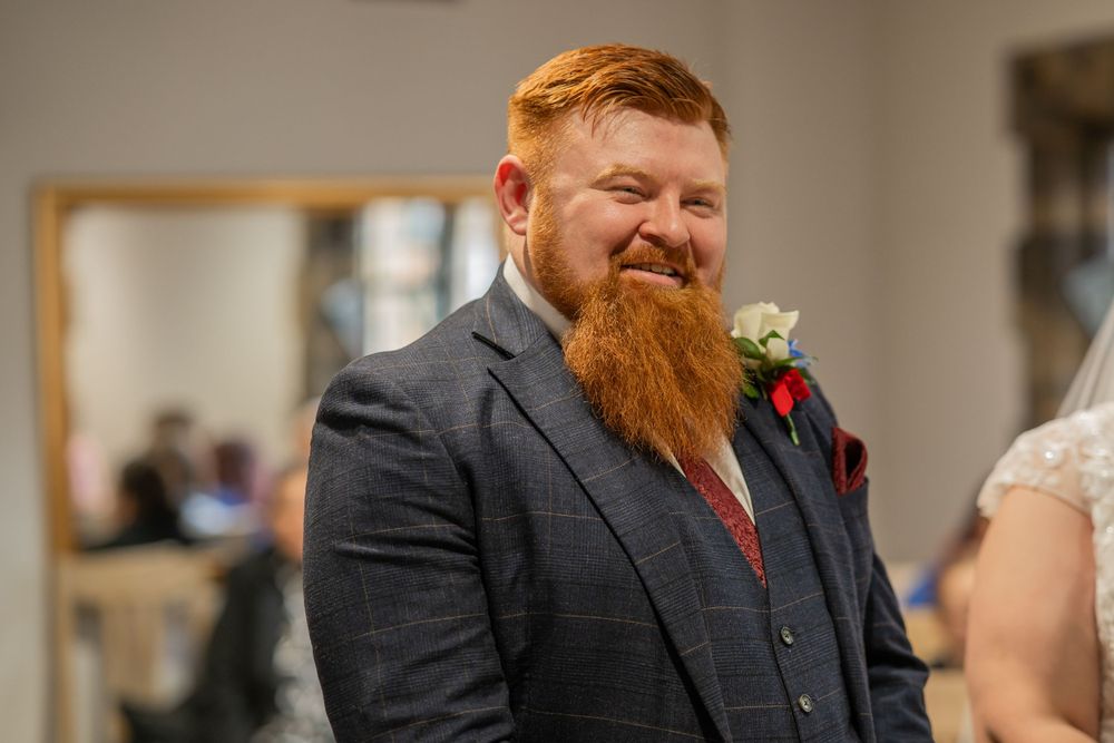 Man with bright red beard wearing gray suit and boutonniere smiles during celebration.