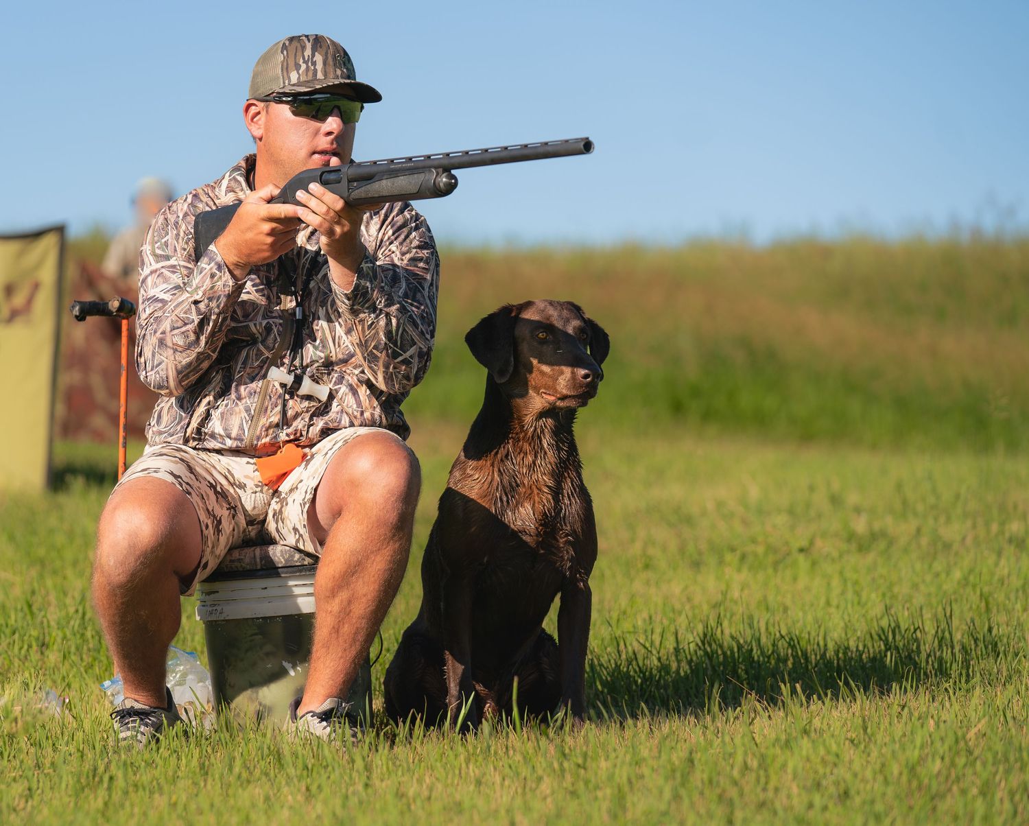 Hunter in camouflage gear sits with hunting dog on lookout post during sunny outdoor hunt.