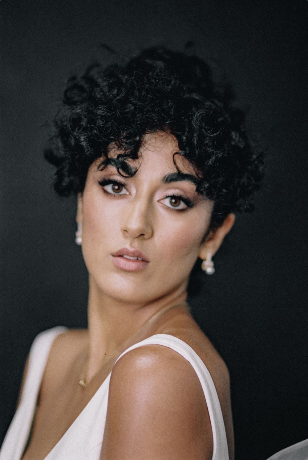 Portrait photo featuring tight curly dark hair, dramatic eye makeup, white sleeveless top, and pearl earrings against dark background.