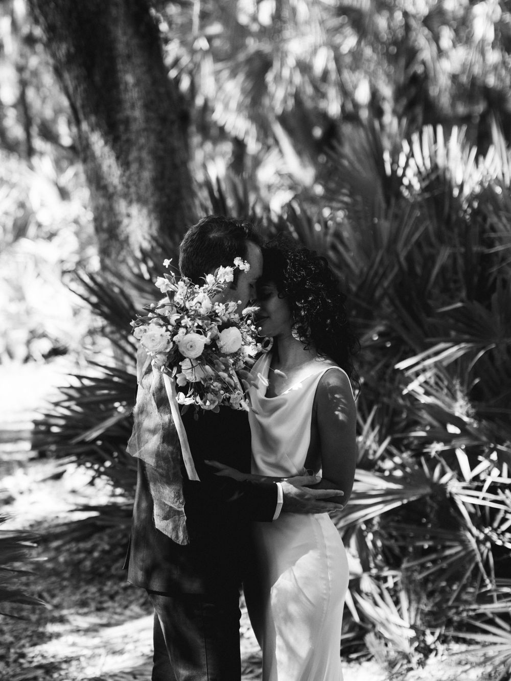 A romantic black and white wedding photo showing a tender embrace among tropical palm fronds and dappled sunlight.