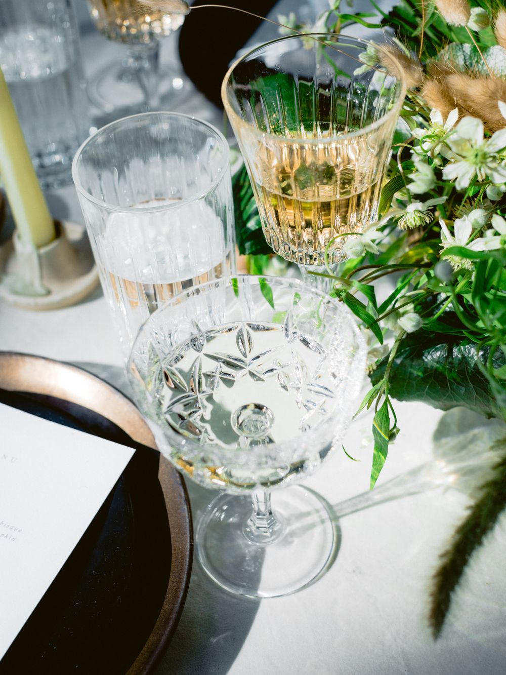 Crystal cocktail glasses with drinks sit elegantly among fresh green foliage and white floral arrangements on a table setting.