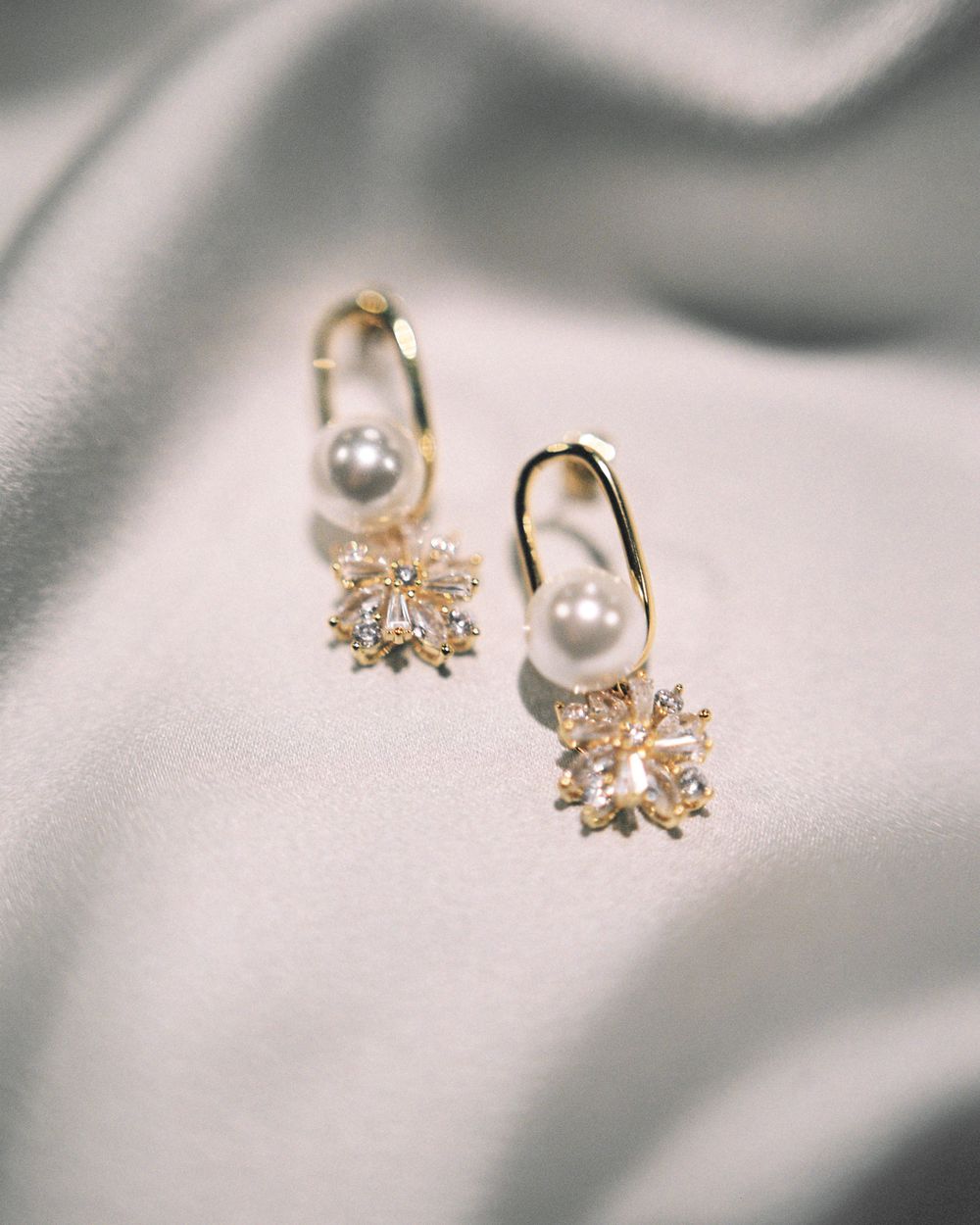 Delicate gold and pearl drop earrings with crystal flower designs photographed on soft gray silk fabric.