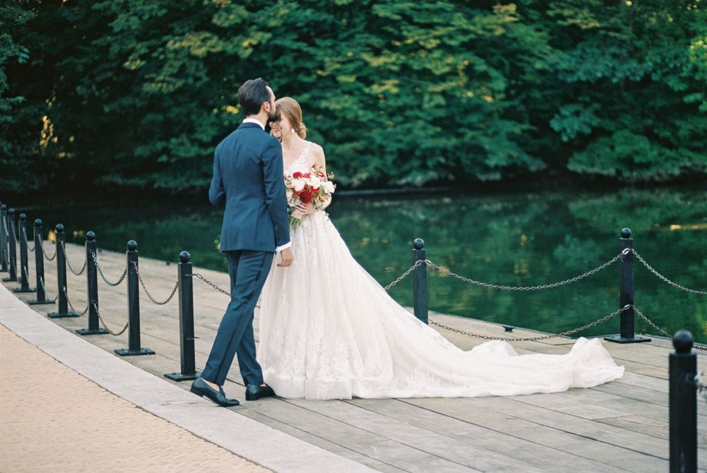 A couple in wedding attire share a tender moment on a railed pathway surrounded by lush green trees.