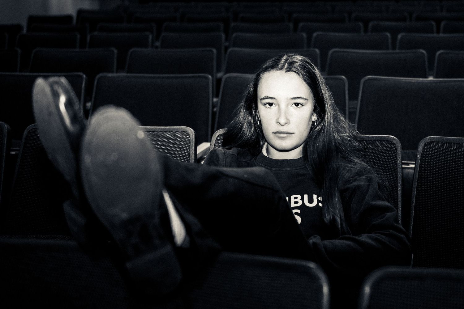 A person in a black sweatshirt sits alone in a dark movie theater with their feet up on the seat in front.