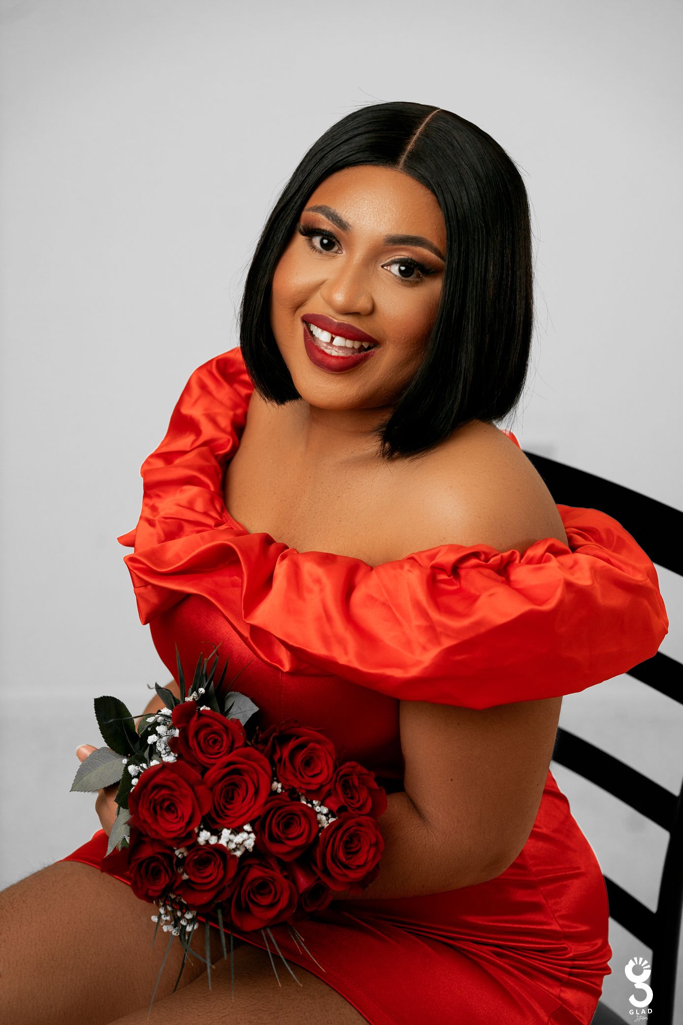 A person in a bright red off-shoulder dress poses with a bouquet of red roses against a light grey background.