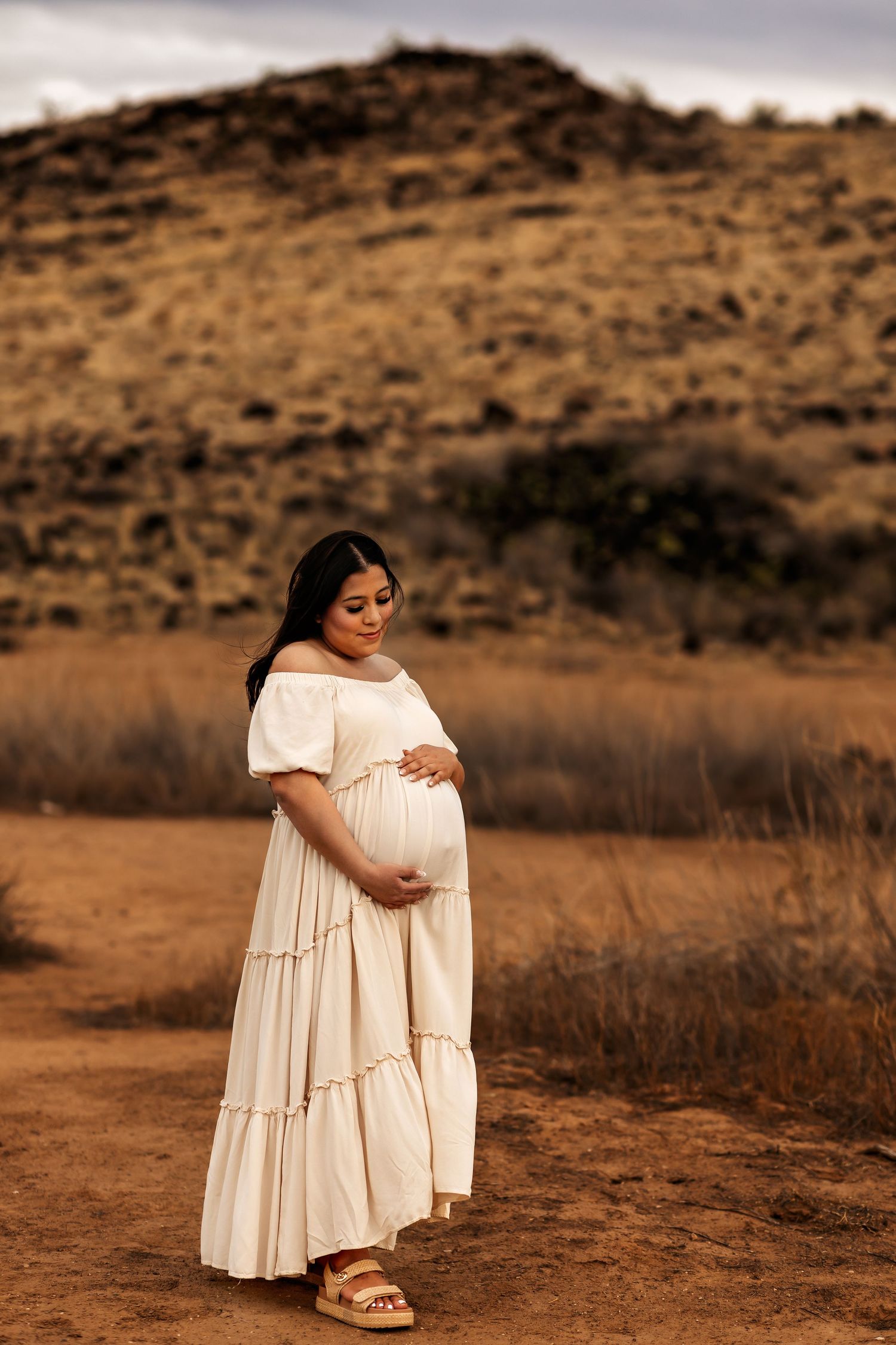 Maternity photo of woman in flowing white dress against desert mountain landscape for her photoshoot.